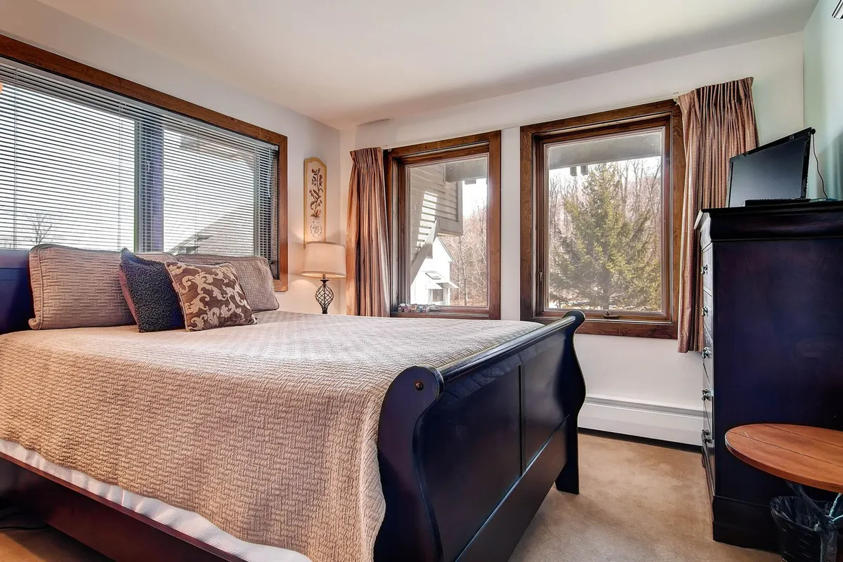 This image presents a cozy bedroom scene featuring a queen-sized bed, inviting linens, a dresser, nightstand with a lamp, and large windows allowing in natural light. The calming ambiance is perfect for relaxation, evoking a sense of comfort and personal retreat.
