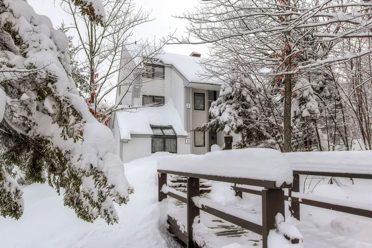 The image captures a snow-covered house set amidst surrounding trees, with a charming wooden bridge in the foreground. The peaceful winter scene hints at outdoor adventure, exploration, and the beauty of the natural environment around Sunrise Mountain Village—ideal for scenic pet walks in all seasons.