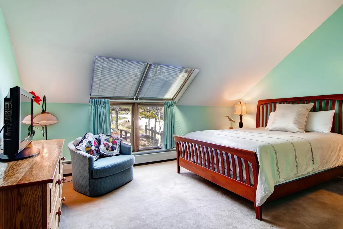 The image showcases a calm and comfortable bedroom with a queen-sized bed, an armchair perfect for reading or unwinding, a dresser, and a window framing peaceful natural views. The decor is unique yet understated, promoting a restful atmosphere.
