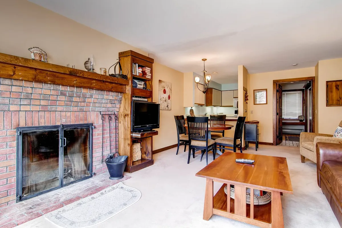 A welcoming living room centered around a classic stone fireplace, surrounded by plush seating and a wooden dining table. The scene is infused with warm light, showcasing both the cozy furniture and a flat-screen TV, creating an inviting space perfect for gatherings. Large windows highlight the room and hint at the seasonal views outside.
