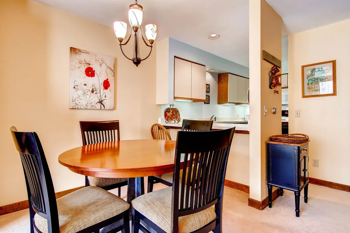 A cozy, inviting dining area featuring a round wooden table under a stylish chandelier. The space is decorated with vibrant wall art depicting red flowers and offers a clear view into the adjacent, modern kitchen. Large windows bring in natural light and frame views of the surrounding forest, enhancing the sense of connection to nature.