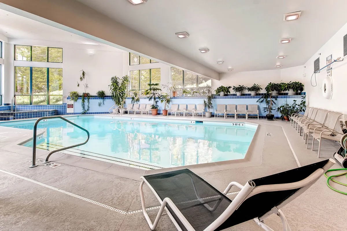 An inviting indoor pool area filled with natural light, featuring sparkling blue water, lounge chairs, potted plants, and large windows offering views of the winter landscape.