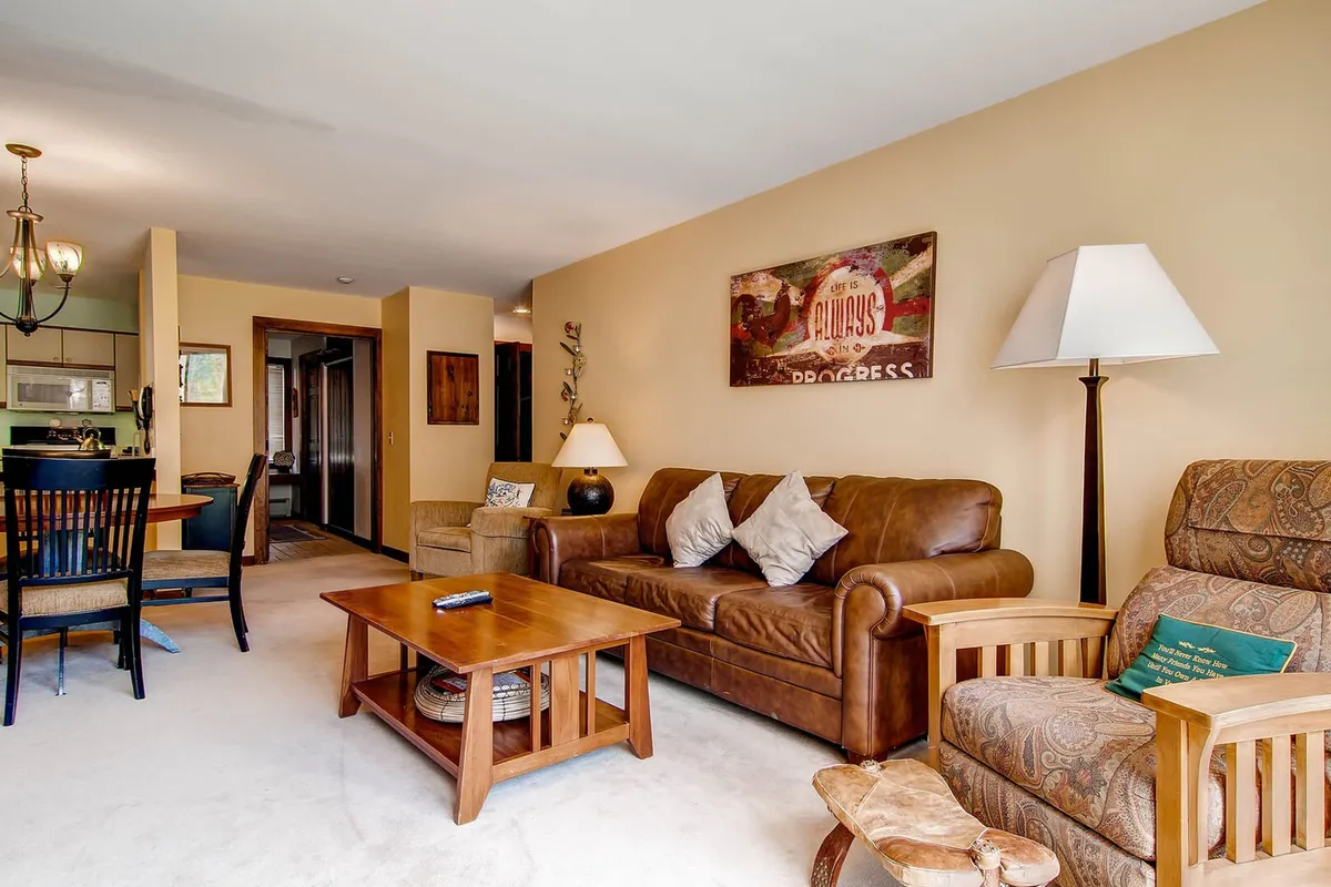 The hero image showcases a warm and inviting living room featuring a plush leather sofa, a rustic wooden coffee table, tasteful decorative wall art, and a gentle floor lamp. The comfortable layout suggests a perfect spot for relaxing after a day on the slopes or gathering with loved ones for cozy movie nights and memorable conversations.