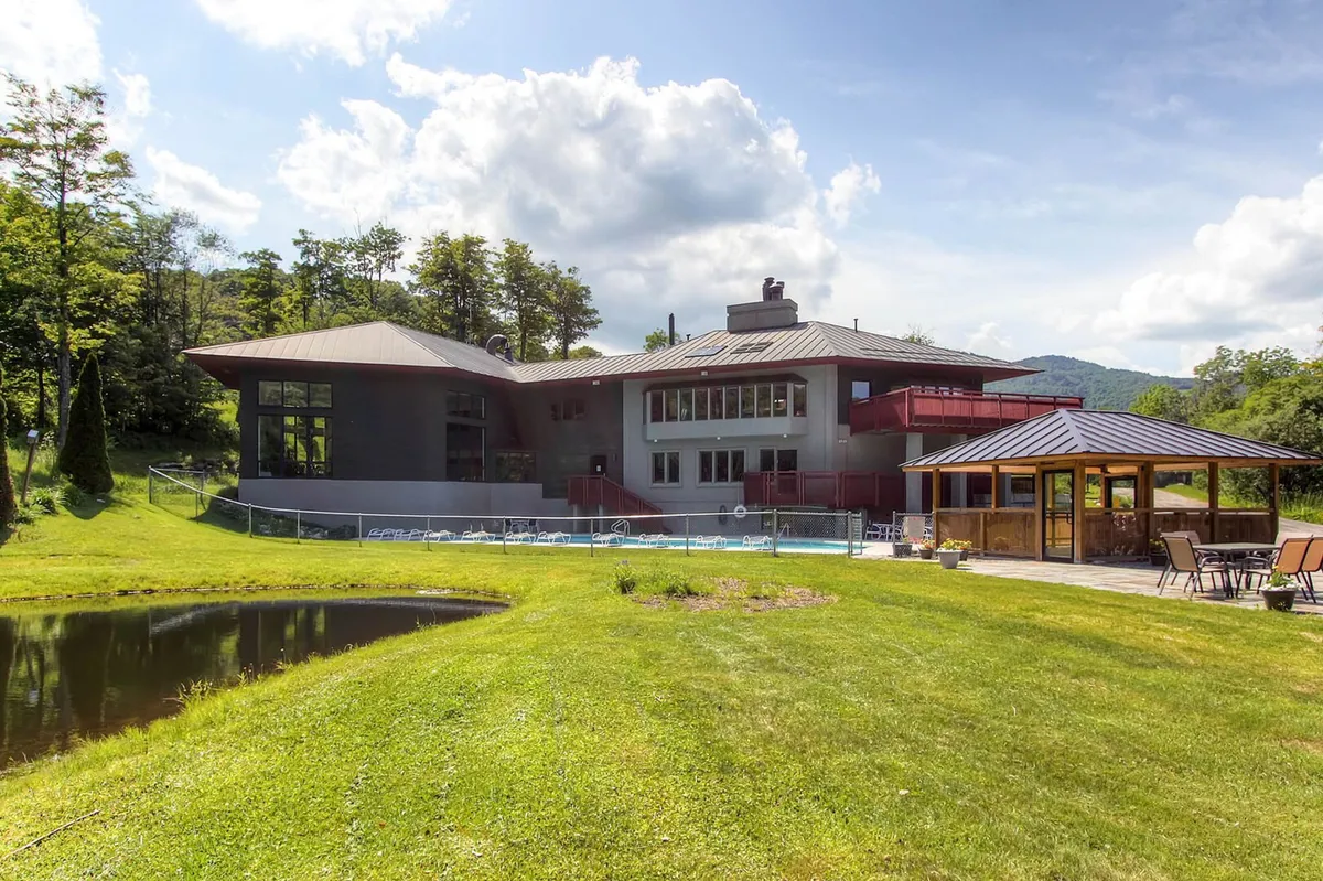 This image features the property's modern architectural design with expansive windows, an outdoor deck area, a reflective pond, and lush surrounding greenery. The deck and pond are set against a tranquil backdrop of mountains and forest, evoking an inviting and peaceful atmosphere ideal for outdoor relaxation and gatherings.